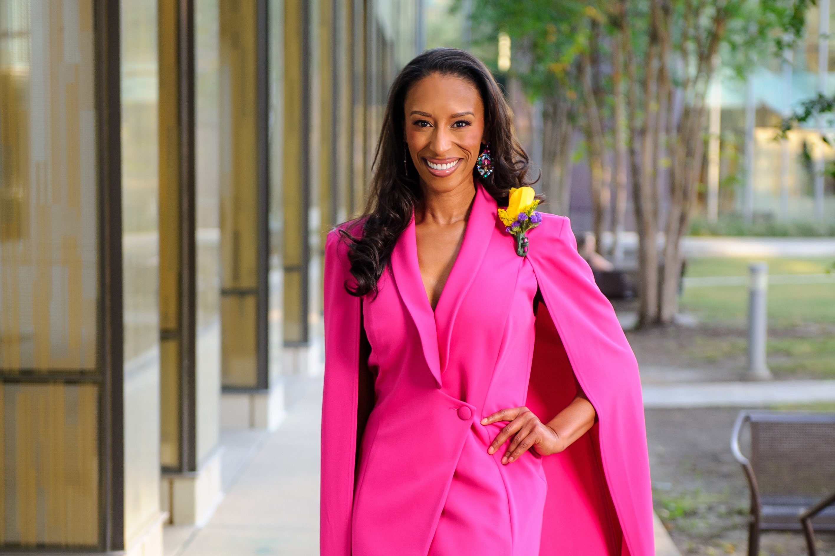 Whitney Breaux is wearing a bright pink dress. She is standing outside with trees and columns in the background. 